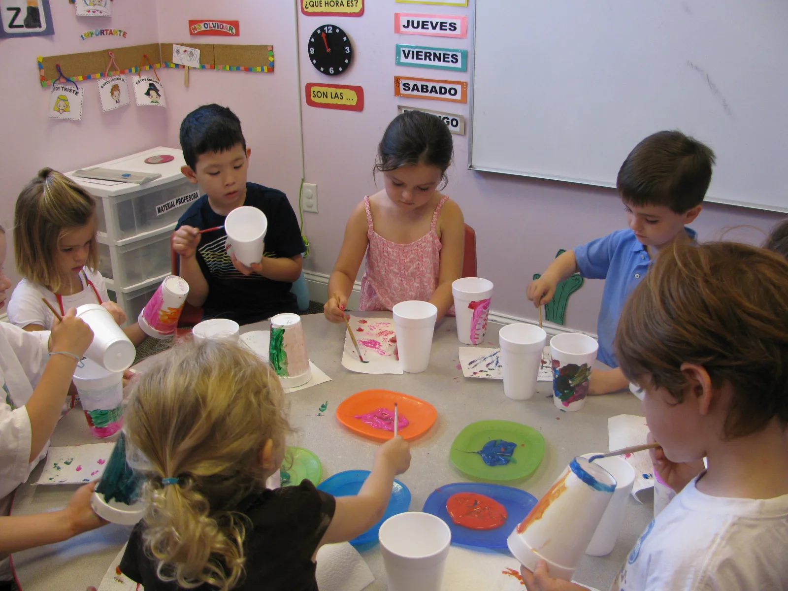 Young SELA students gather around a classroom table for a group painting activity, with Spanish day-of-the-week cards displayed on the wall behind them.