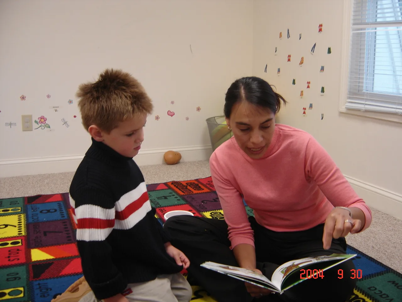 A SELA teacher and a young student sit together on a letters-and-numbers rug, reading a picture book.