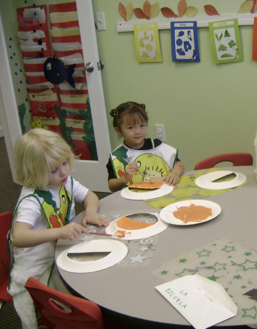 Two young SELA students in art smocks paint together at a classroom table, with Spanish color words posted on the wall behind them.