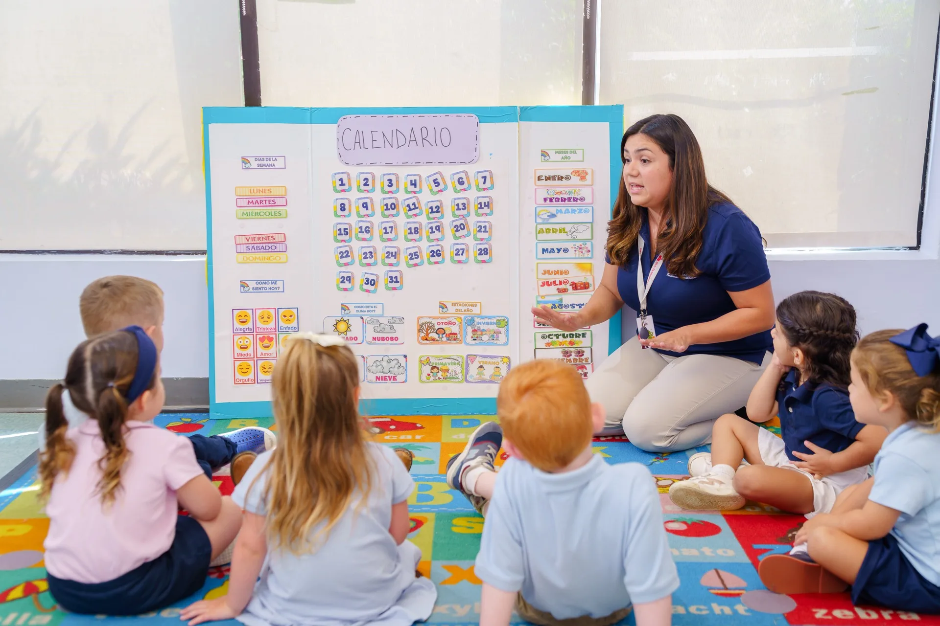 A SELA teacher kneels at a Spanish ‘Calendario’ board, leading a circle of young children in a language lesson.