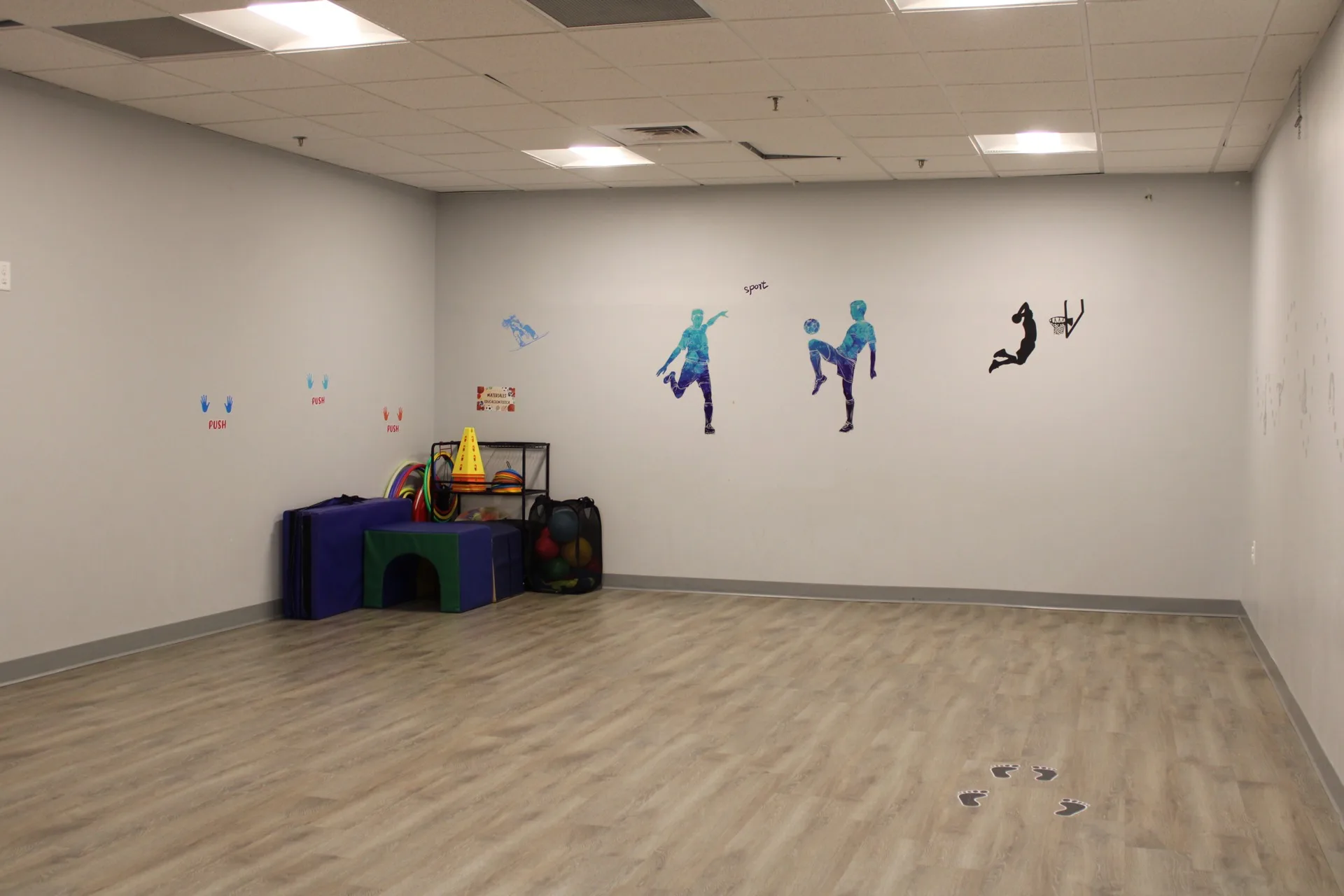 An indoor movement room at SELA's Hingham campus, with an open wood floor, soft-play equipment, and sport wall art.
