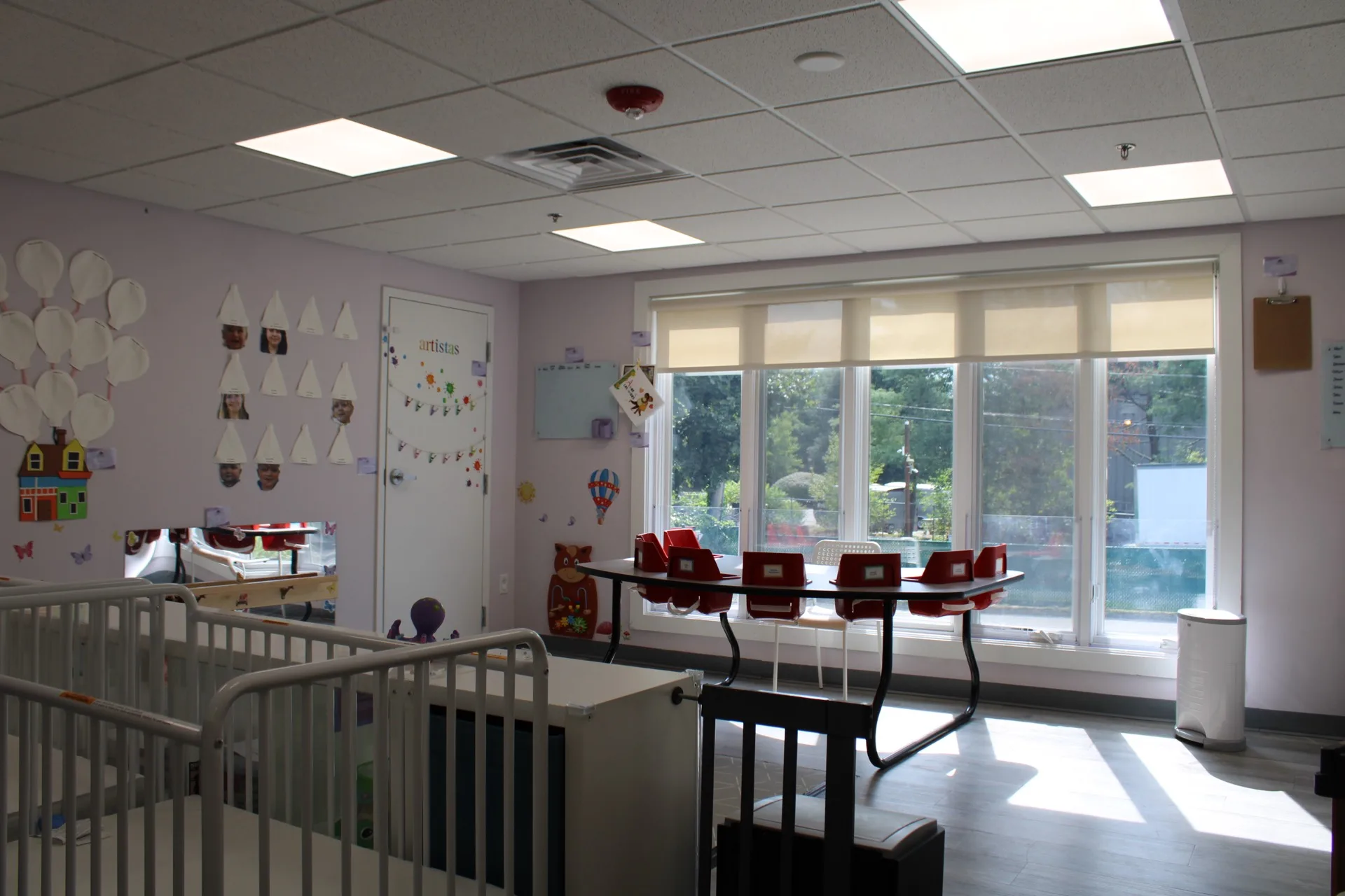 An early-years classroom at SELA's Norwell campus, with cribs and a sunlit activity table by large windows.