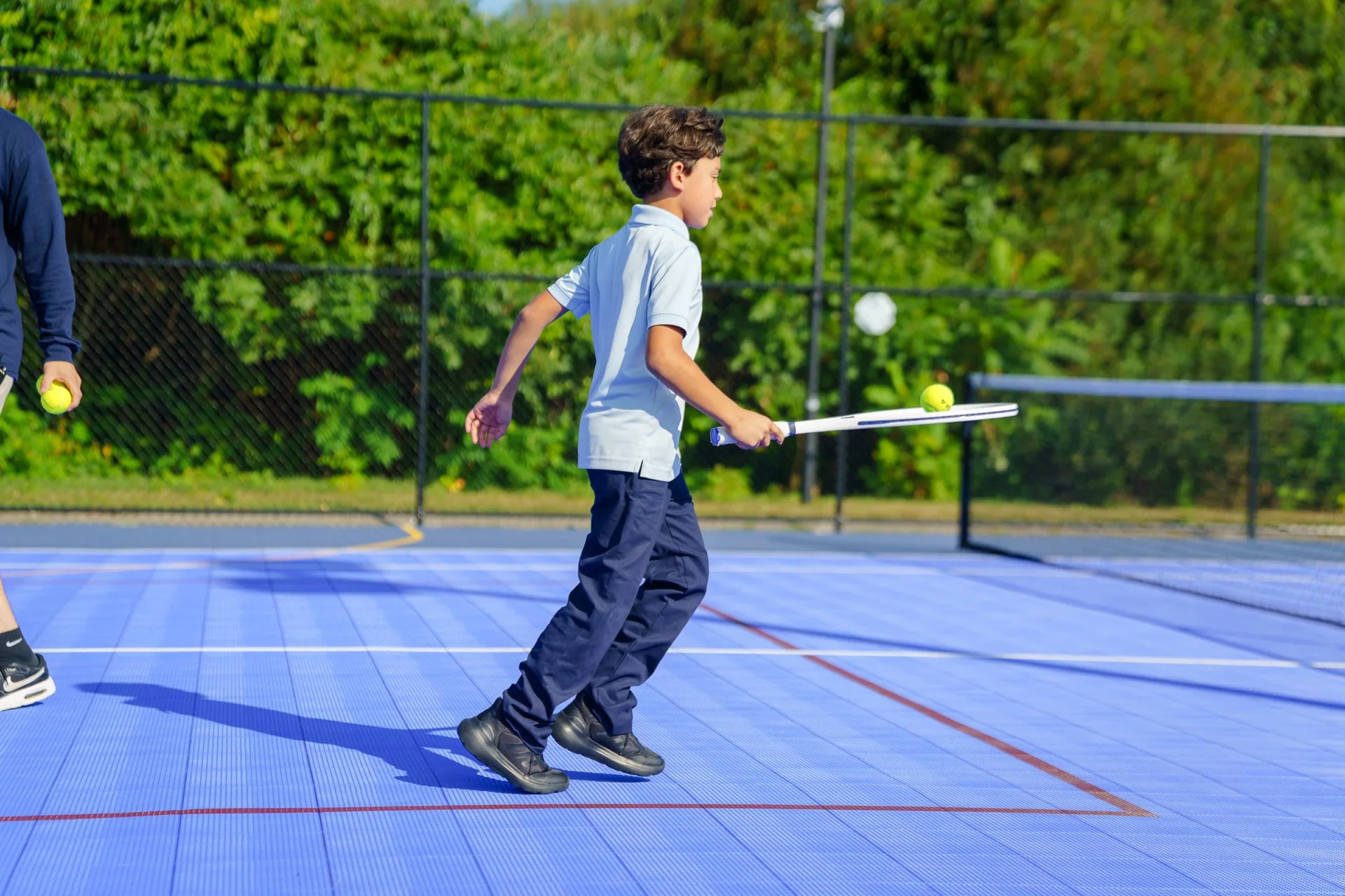 A SELA child swings a tennis racket mid-stride on the blue outdoor court.