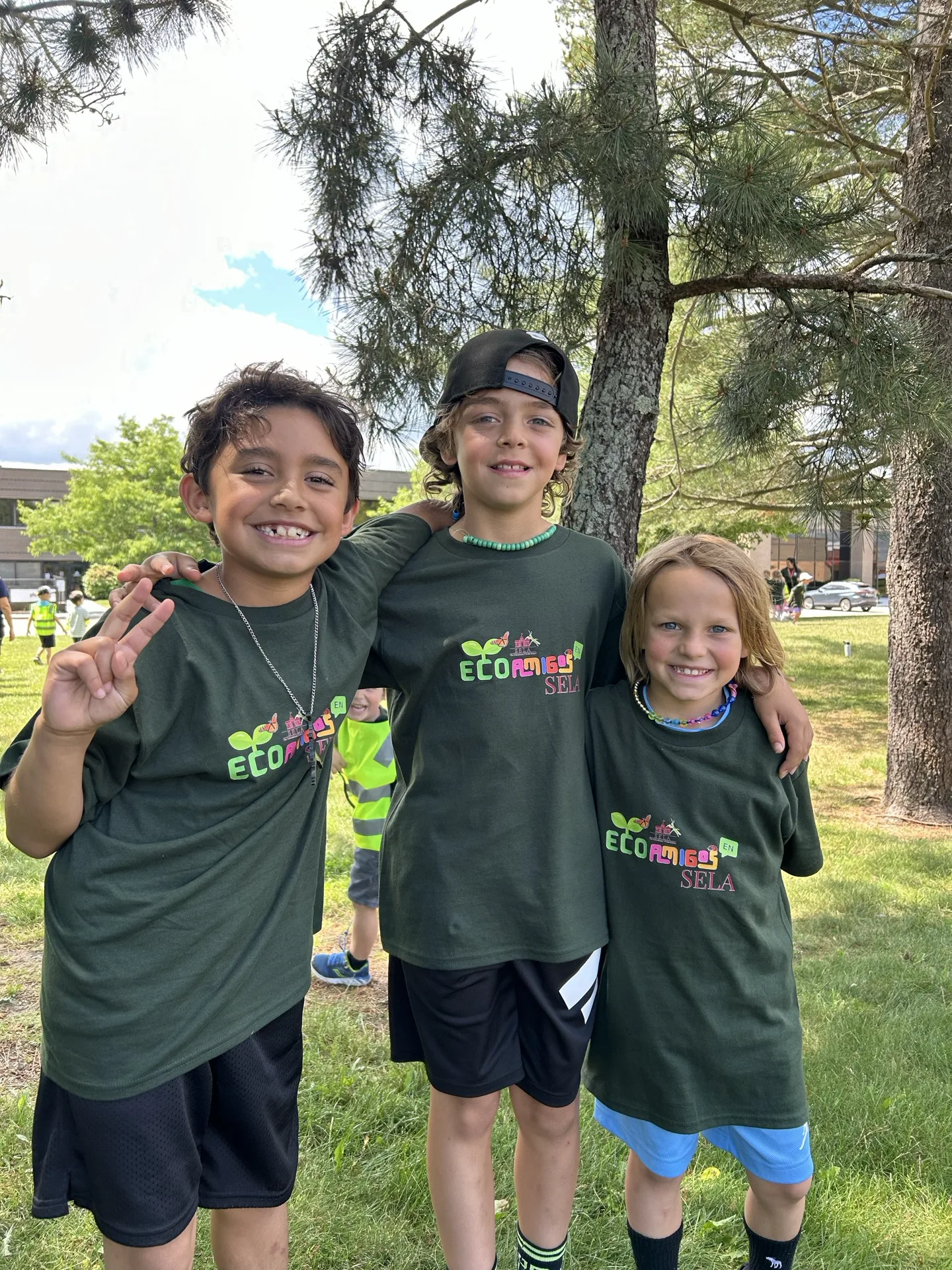 Three SELA campers in green Eco Amigos t-shirts pose arm-in-arm under the trees on a summer afternoon.