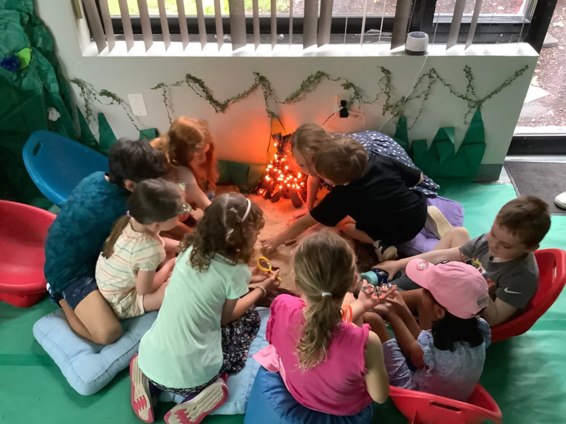 SELA children gather close around a pretend indoor campfire of lights and logs during a summer story circle.