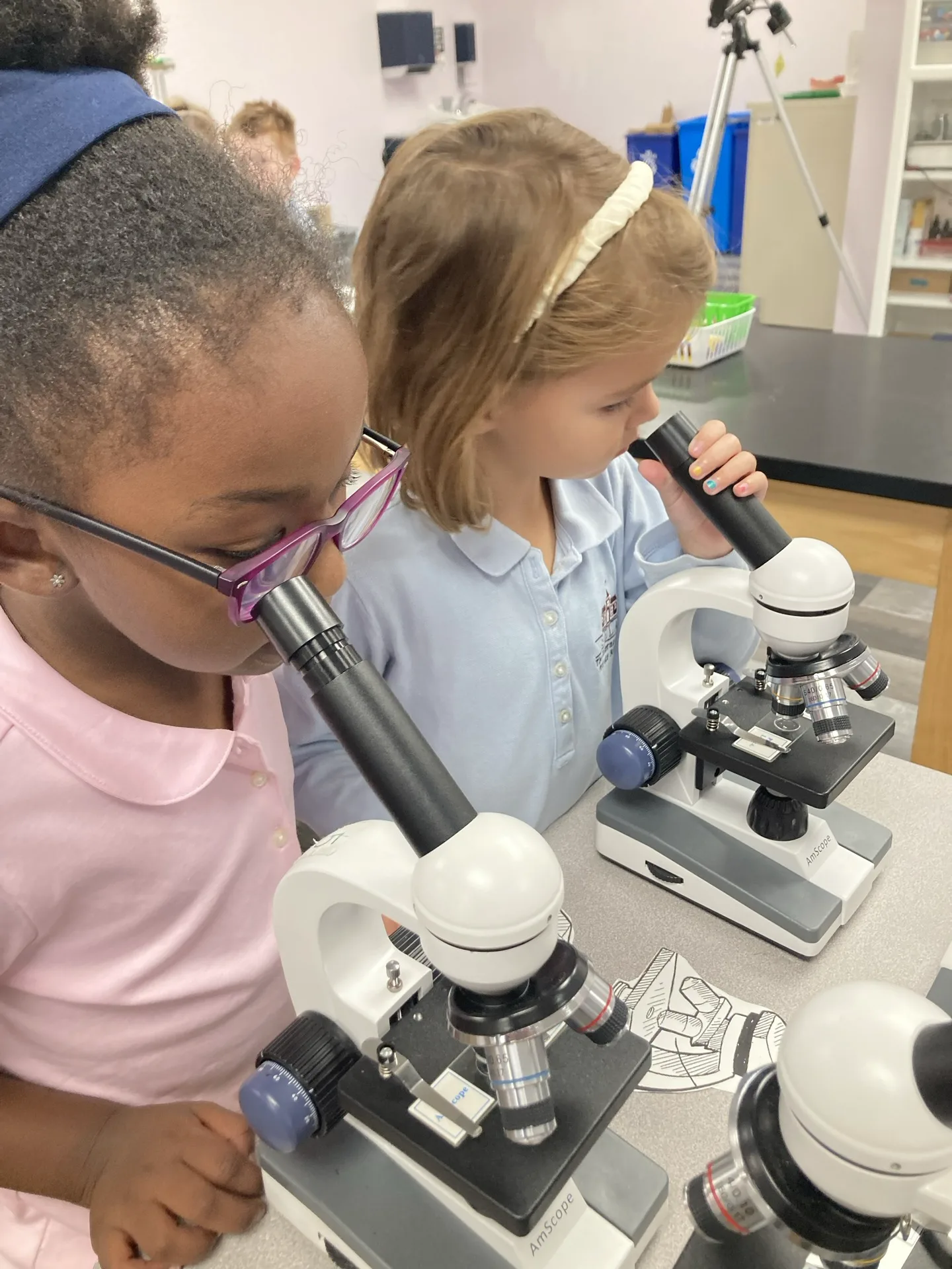 Two SELA Lower School girls lean into microscopes side by side during a science lesson.