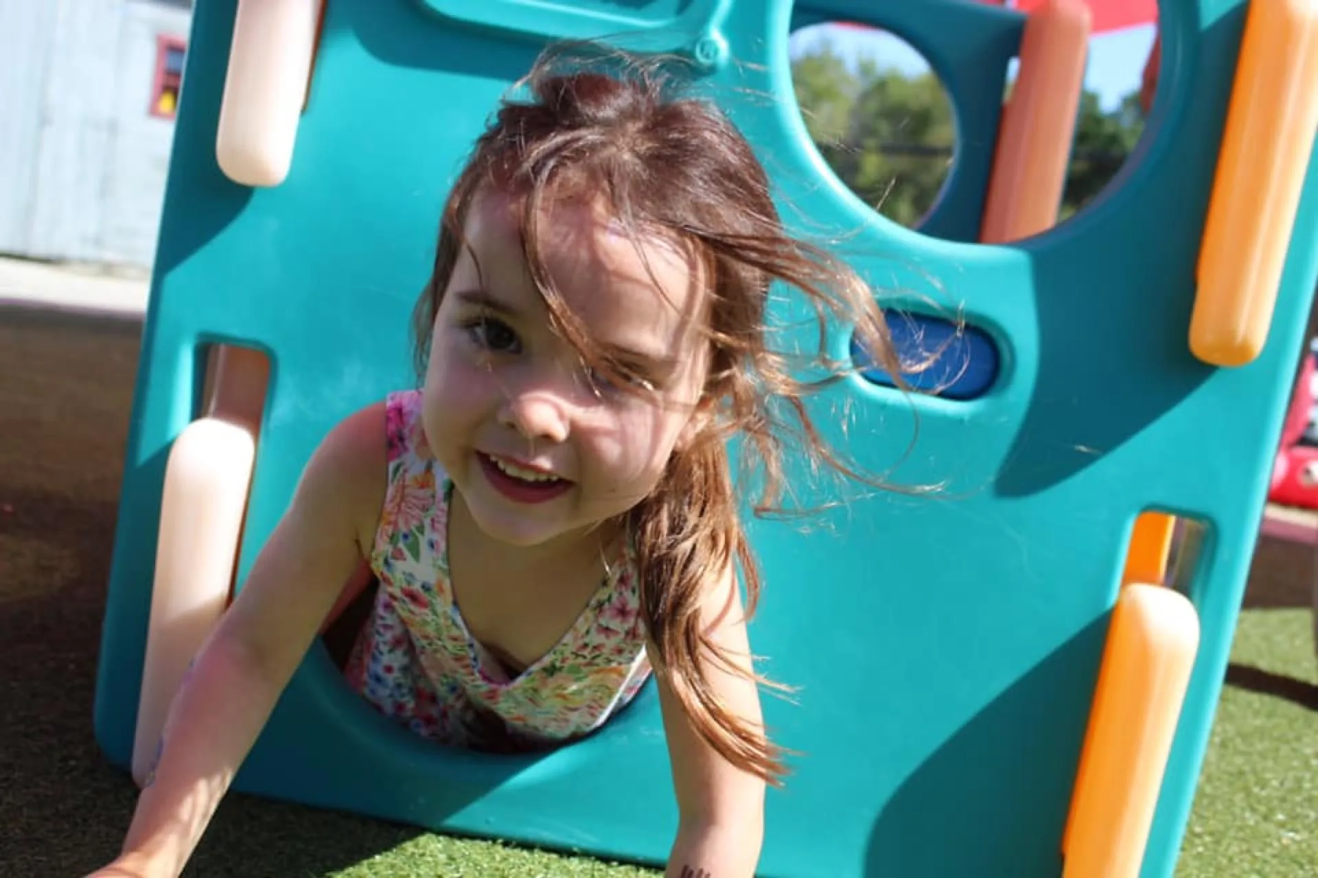 A SELA child grins as she crawls out of a teal playground tunnel, hair caught mid-motion in the wind.